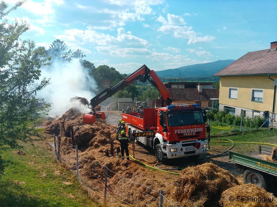Ein Feuerwehrfahrzeug mit ausgestrecktem Kran löscht einen Brand in einem Heuhaufen. Zwei Feuerwehrleute in gelber und schwarzer Uniform stehen in der Nähe.