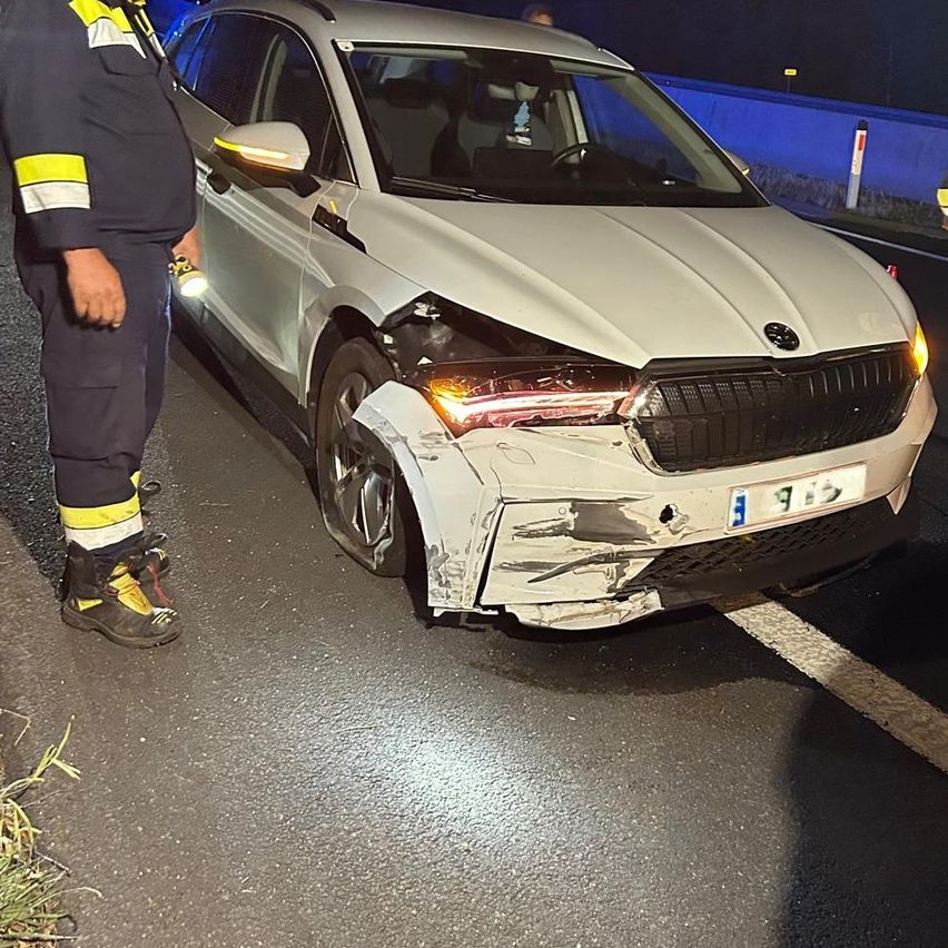 Ein weißer Wagen mit beschädigtem vorderen Stoßfänger und Scheinwerfern steht nachts am Straßenrand. Eine Person in einer reflektierenden Weste steht in der Nähe.