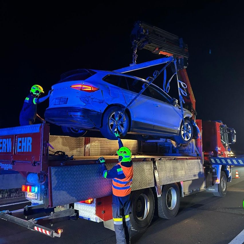Zwei Arbeiter in Schutzausrüstung heben ein beschädigtes Auto auf einen Pritschenwagen, bei Nacht. Das Auto ist auf dem Kopf, und der LKW hat einen roten Körper mit dem Wort 'EHR' an der Seite.