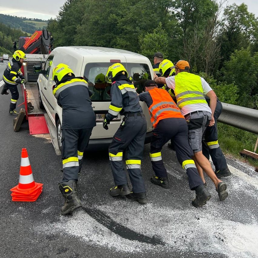 Rettungskräfte in Warnwesten schieben einen weißen Lieferwagen von der Straße. Sie befinden sich am Straßenrand mit Verkehrskegeln und einem Lastwagen im Hintergrund.