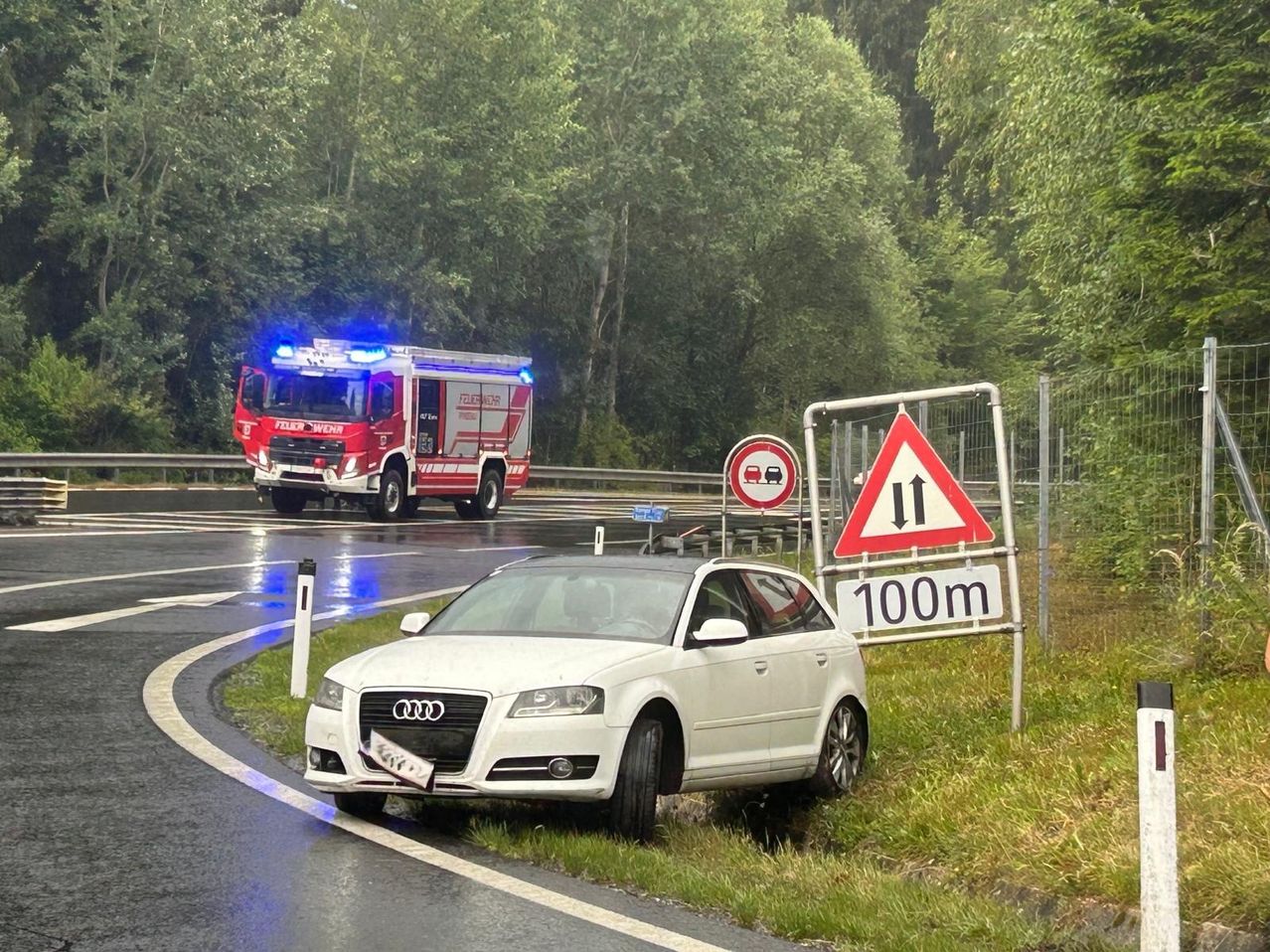 Ein weißer Audi-Wagen ist am nassen Straßenrand geparkt, mit einem Feuerwehrwagen im Hintergrund. Ein dreieckiges Schild mit einem nach oben zeigenden Pfeil und der Nummer 100 ist sichtbar.