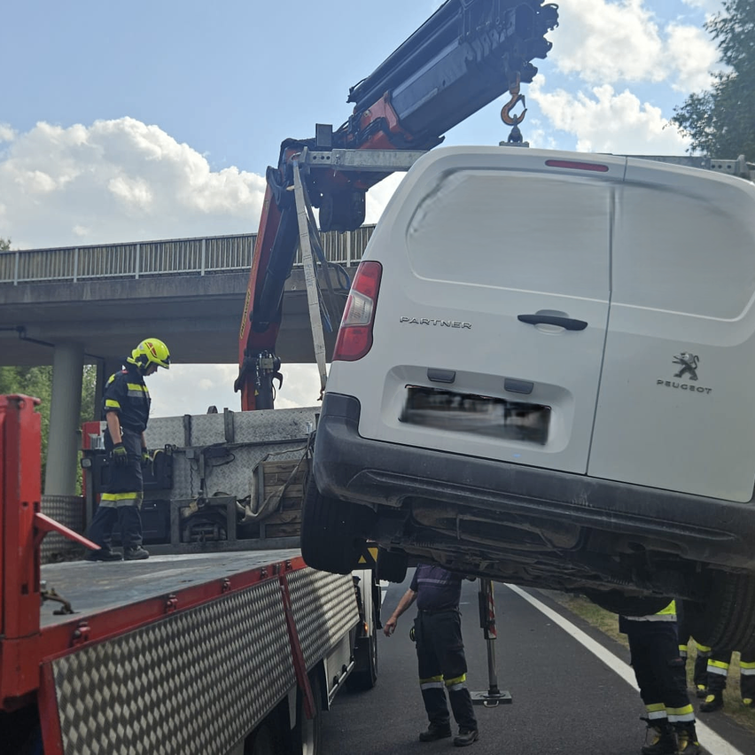 Ein weißer Peugeot-Transporter wird von einem Kran auf die Ladefläche eines roten Lastwagens gehoben. Eine Person in einem gelben Helm steht auf der Ladefläche.