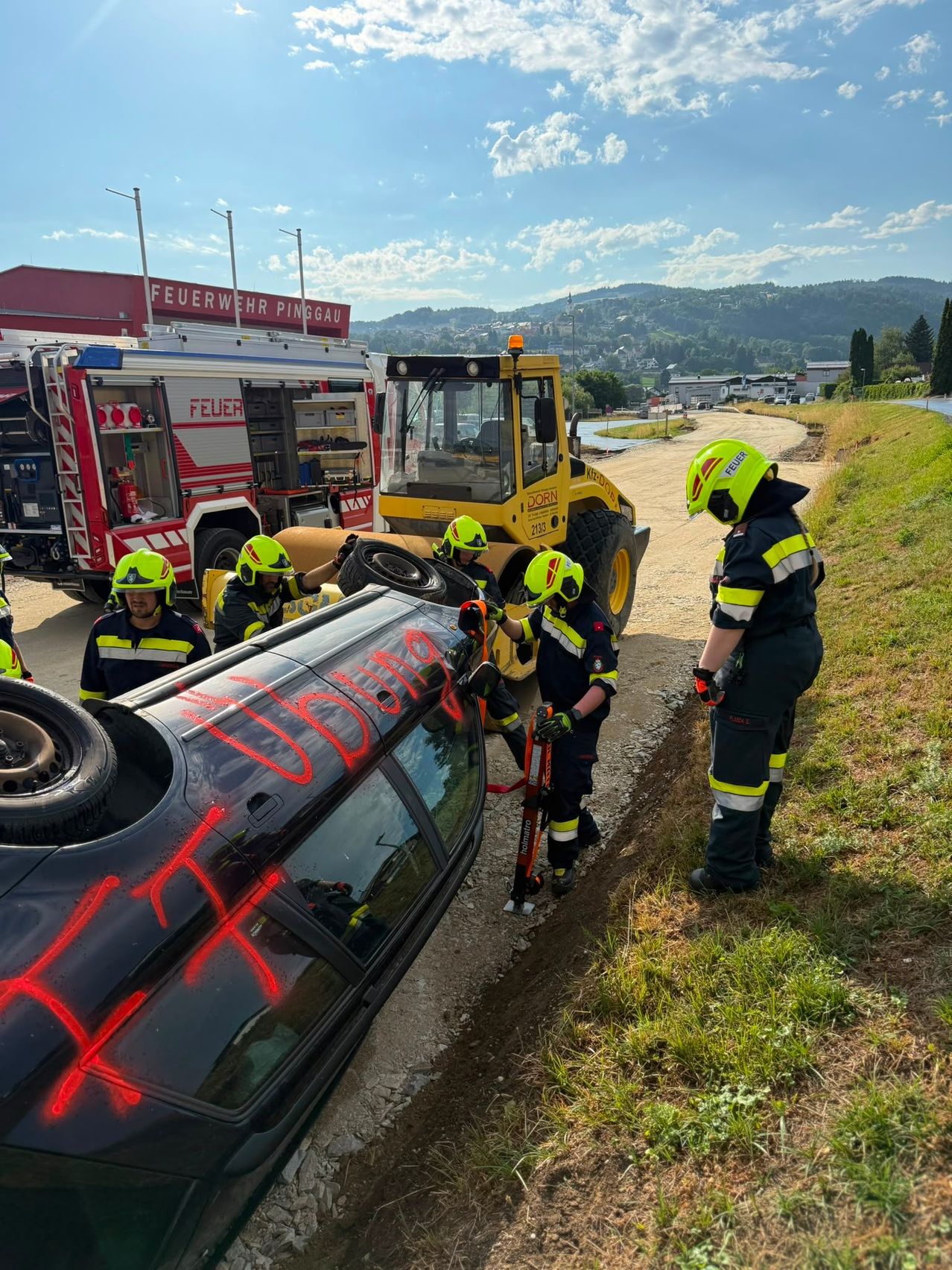 Eine Gruppe Feuerwehrleute in gelben und grünen Helmen arbeitet an einem umgekippten schwarzen Auto mit rotem Graffiti am Straßenrand. Ein Feuerwehrwagen und ein gelber Bulldozer sind in der Nähe geparkt.