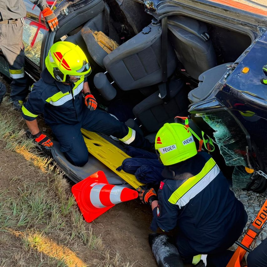 Rettungskräfte in gelben Helmen arbeiten daran, eine Person aus einem beschädigten Fahrzeug auf einem Grasfeld zu extrahieren. Ein Verkehrskegel und eine zerbrochene Windschutzscheibe sind sichtbar.