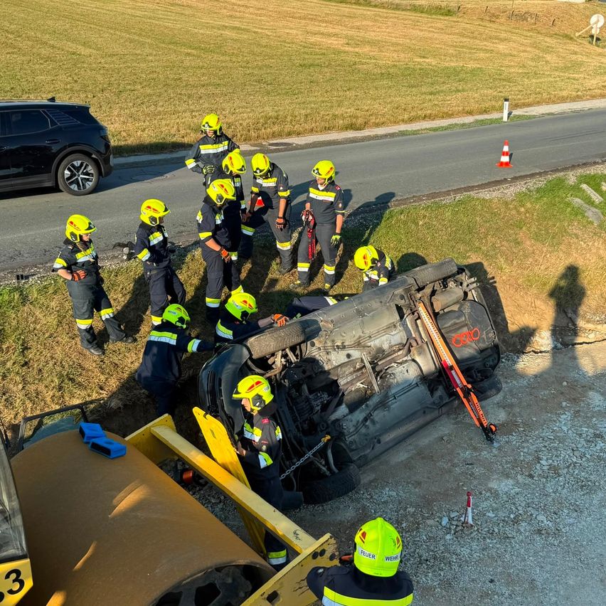 Eine Gruppe von Feuerwehrleuten in gelben Helmen arbeitet an einem umgekippten Auto am Straßenrand.