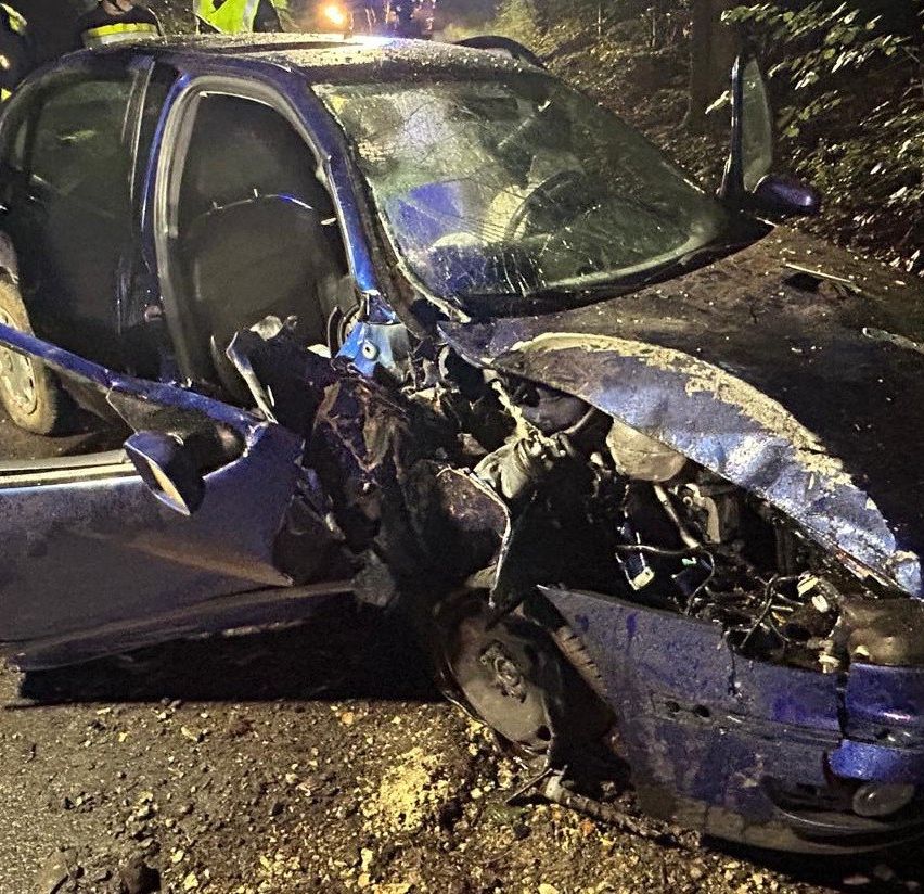Ein schwer beschädigtes blaues Auto ist zu sehen, dessen Front vernichtet ist und die Fahrertür offen steht. Rettungskräfte sind anwesend.