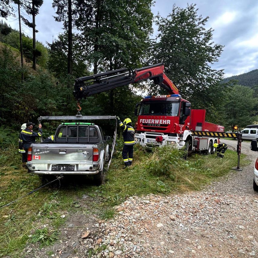 Ein roter Feuerwehrwagen benutzt einen Kran, um einen silbernen Pickup-Truck vom Boden zu heben. Zwei Feuerwehrleute stehen in der Nähe, und ein weiterer ist am Boden.