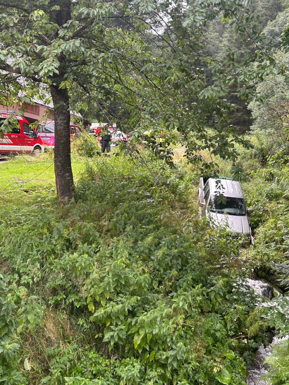 Ein weißer SUV ist in einem Waldgebiet steckengeblieben, umgeben von dichter Vegetation. In der Nähe sind Rettungsfahrzeuge geparkt, und mehrere Personen stehen um das Fahrzeug herum.