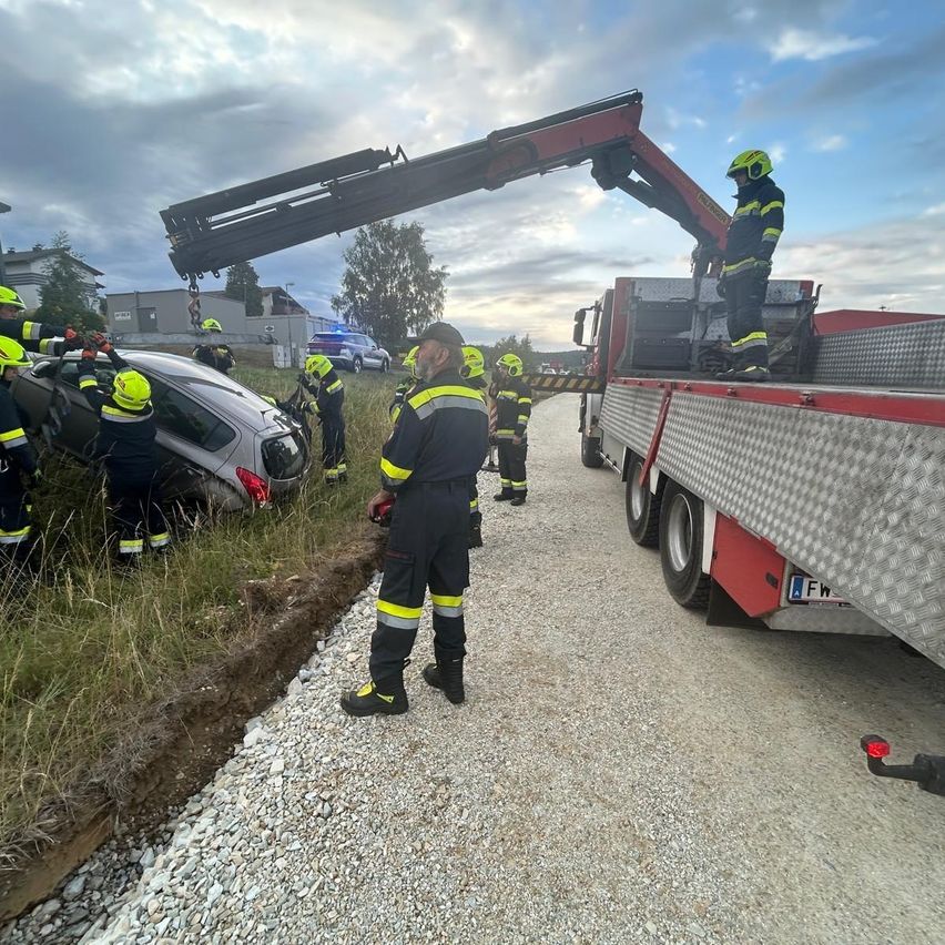 Feuerwehrleute benutzen einen Kran, um ein beschädigtes Auto von der Straße zu heben. Mehrere Personen in gelben Helmen helfen.