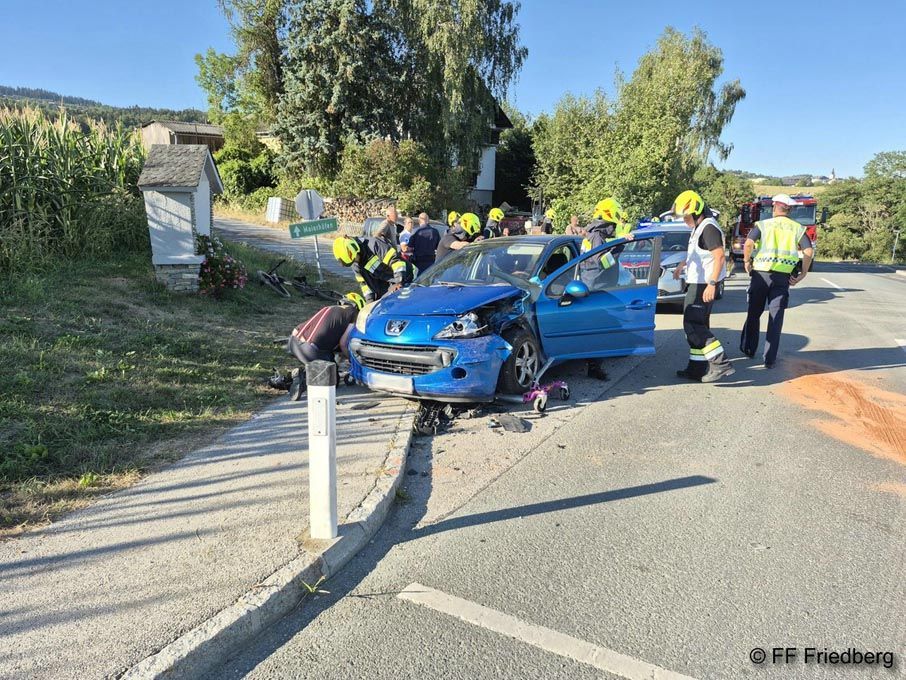Ein blaues Auto ist auf der Straßenseite in einen Pfosten gecrasht. Rettungskräfte in gelben Helmen sind am Unfallort.