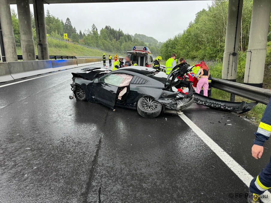 A black sports car is wrecked on the side of a road. Emergency personnel attend to the scene, with one person in the car.