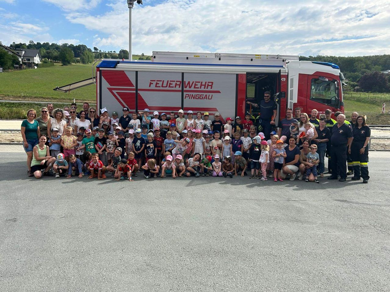 A group of children and adults pose for a photo in front of a red fire truck labeled 'Feuerwehr Pingggau'.
