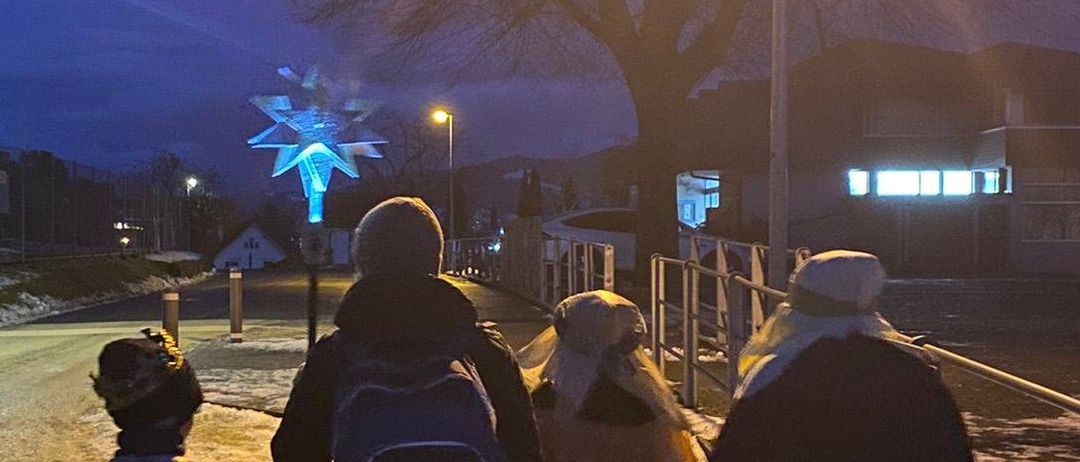 Vier Personen gehen bei Nacht einen verschneiten Straßenweg entlang, beleuchtet von Straßenlaternen. Ein Stern ist über ihnen sichtbar.