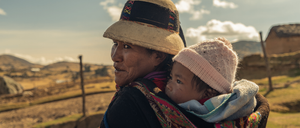 Eine Frau mit einem breitkrempigen Hut und einem bunten Schal trägt ein Baby in einer Trage. Sie stehen auf einer grasbewachsenen Anhöhe mit einem verschwommenen Bergrücken im Hintergrund.