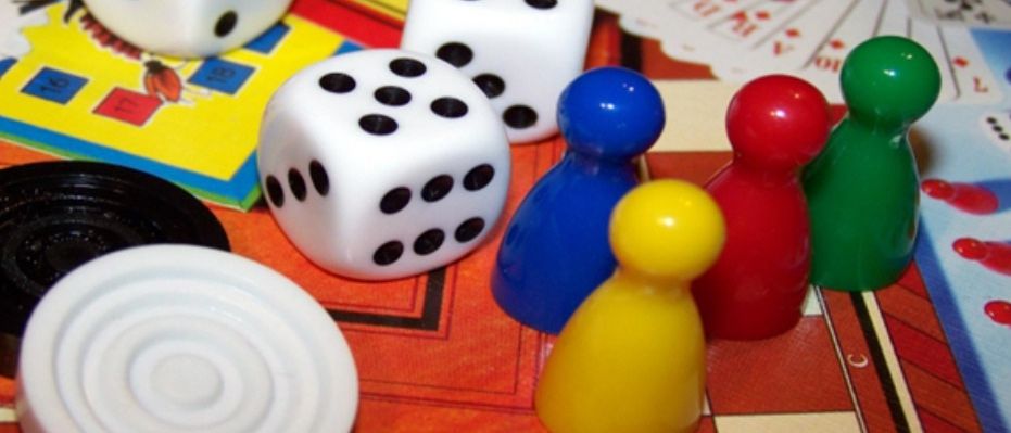 A tabletop game setup with dice, playing cards, and colorful pawns. The game board has a checkered pattern.
