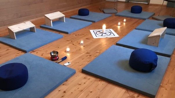 A serene room with wooden floors, several blue cushions, and meditation benches. A lit candle is placed in the center on a diagram. Other objects, like a bowl and a phone, are scattered around.