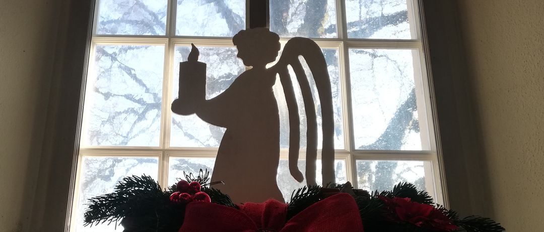 Ein Fenstersims mit einer Weihnachtsdekoration, bestehend aus einer Silhouette eines Engels, der eine brennende Kerze hält, umgeben von roten Blumen und Grün.
