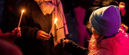 Eine Person hält zwei brennende Kerzen und steht mit einem Kind im Dunkeln. Das Kind trägt eine Mütze und einen Schal.