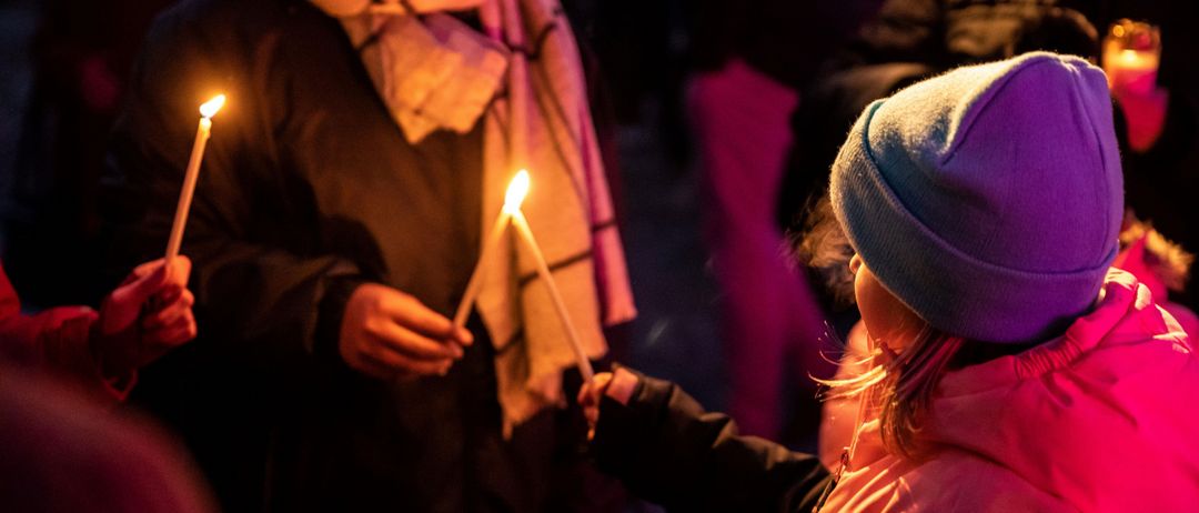 Eine Person hält zwei brennende Kerzen und steht mit einem Kind im Dunkeln. Das Kind trägt eine Mütze und einen Schal.