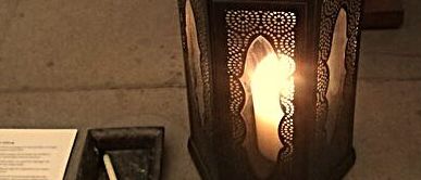 Eine Laterne mit einer brennenden Kerze steht auf einem Tisch. Daneben befindet sich ein kleiner Teller mit einer einzelnen Kerze. Ein Stück Papier mit Text liegt ebenfalls auf dem Tisch.