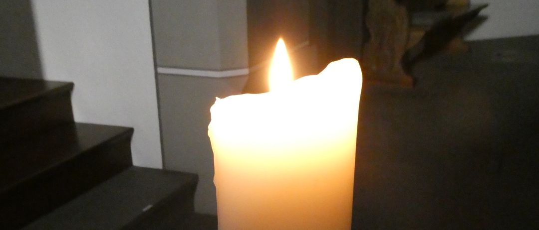 Eine Kerze in einem schwach beleuchteten Raum. Sie hat ein Design einer Taube und einer Sonne auf der Seite. Es gibt Holzbänke und Treppen.