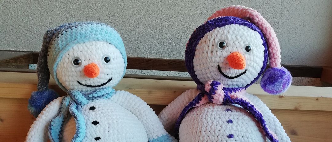 Zwei gehäkelte Schneemänner sitzen nebeneinander auf Holzstufen. Beide tragen Mützen, Schals und haben orange Nasen.