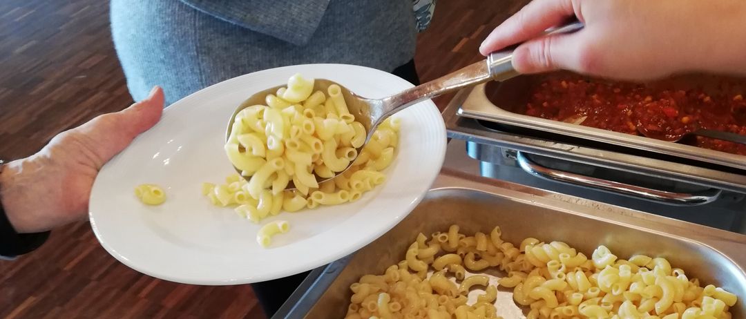 Zwei Personen servieren Mac and Cheese von einer Buffetlinie; eine Person hat einen vollen Teller, während eine andere mehr auf ihren Teller löffelt.
