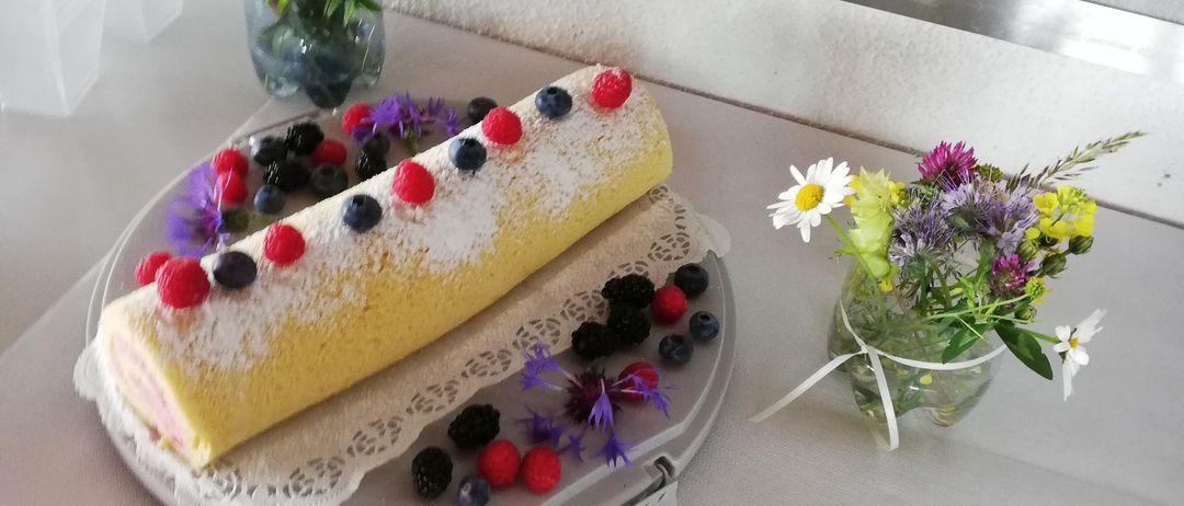 Ein Teller mit einem gerollten Kuchen, der mit Beeren und Blumen garniert ist, steht auf einer Theke. Dahinter hält ein Regal Stapel von Tellern und eine Box mit Besteck.