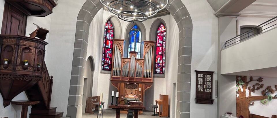Innenansicht einer Kirche mit einem hölzernen Altar, Kirchenbänken und einer Orgel. Eine runde Kronleuchter hängt von der Decke, und eine Blumengestecke ist auf dem Altar. Die Kirche hat Buntglasfenster und eine hölzerne Kanzel.