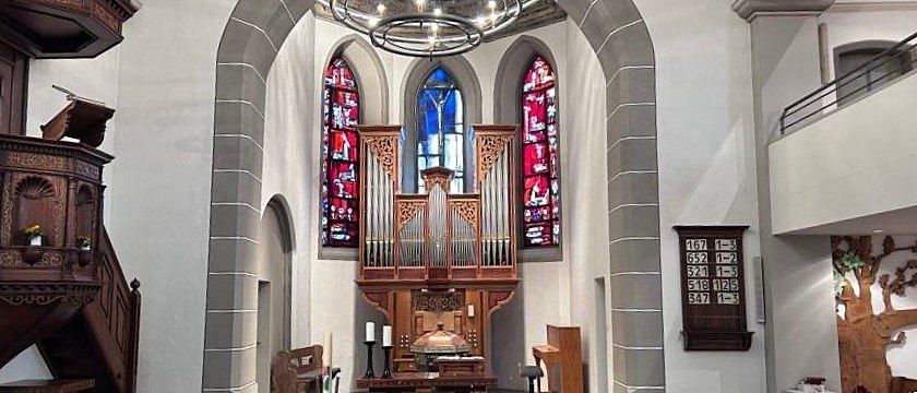 Ein Kircheninneres mit einem hölzernen Altar, einer großen Orgel und Buntglasfenstern. Eine Kronleuchter hängt von der Decke, und eine hölzerne Tafel mit Zahlen ist an der Wand montiert.