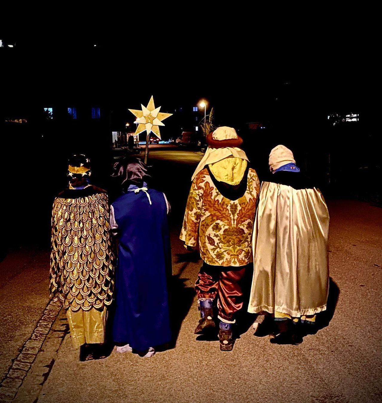 Vier Personen in Kostümen, möglicherweise für ein Fest, gehen bei Nacht auf einer Straße mit einem Stern und einer Straßenlaterne im Hintergrund.