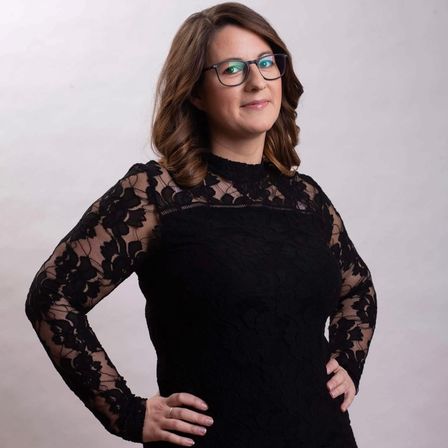 Eine Frau mit braunem Haar, in einem schwarzen Spitzenkleid, mit Brille und Ring, posiert selbstbewusst mit einer Hand auf der Hüfte.