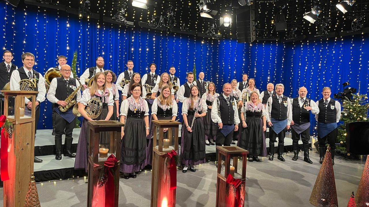 Eine Gruppe von Musikern, sowohl Männer als auch Frauen, posiert für ein Foto auf einer Bühne und hält Musikinstrumente in den Händen. Sie tragen traditionelle Kleidung und stehen hinter brennenden Kerzen.