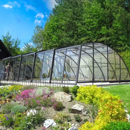 Ein Gewächshaus mit Glasdach und schwarzem Rahmen steht in einem Garten mit verschiedenen Pflanzen und Bäumen. Eine Person steht im Inneren des Gewächshauses.