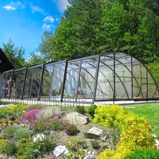 Ein Gewächshaus mit Glasdach und schwarzem Rahmen steht in einem Garten mit verschiedenen Pflanzen und Bäumen. Eine Person steht im Inneren des Gewächshauses.