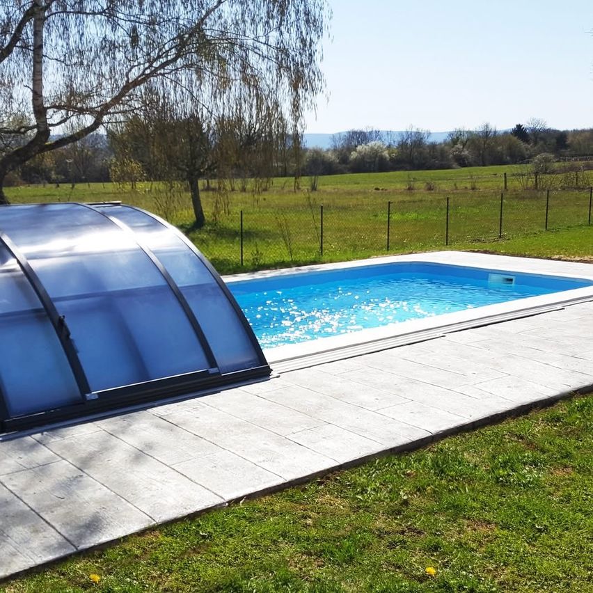 Ein Freibad mit Glasüberdachung befindet sich auf einer Wiese mit einem Baum im Hintergrund unter einem klaren Himmel.
