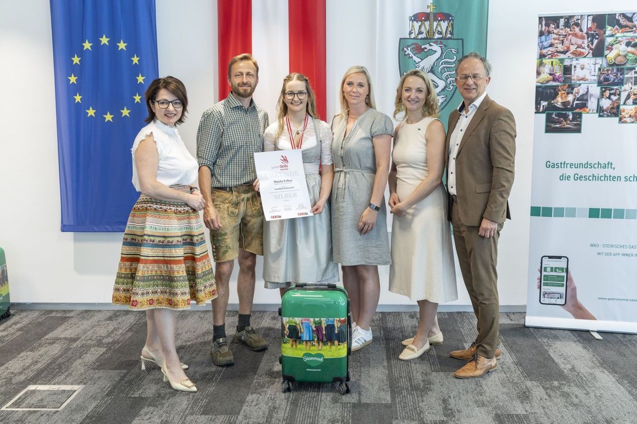 Eine Gruppe von sechs Personen steht zusammen, eine hält ein Zertifikat. Flaggen aus Deutschland und der EU sind dahinter. Ein grüner Koffer steht davor.