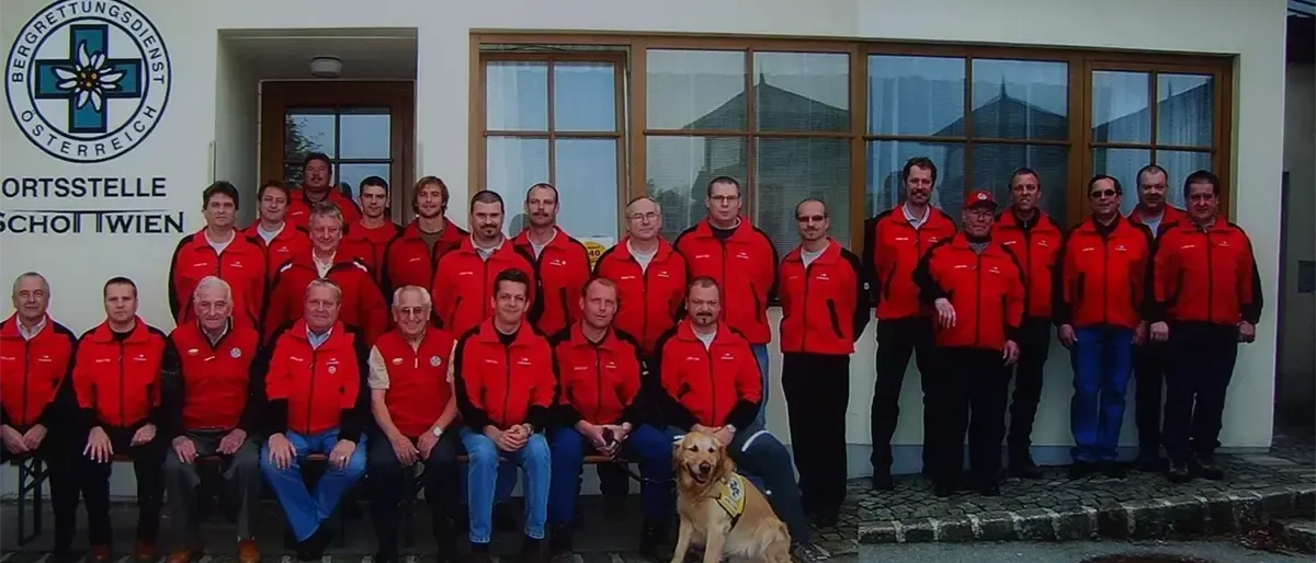 Eine Gruppe von Menschen in roten Jacken, einige sitzend und einige stehend, posiert für ein Foto vor einem Gebäude. Ein Hund mit einem gelben Ausweis sitzt in der Mitte.