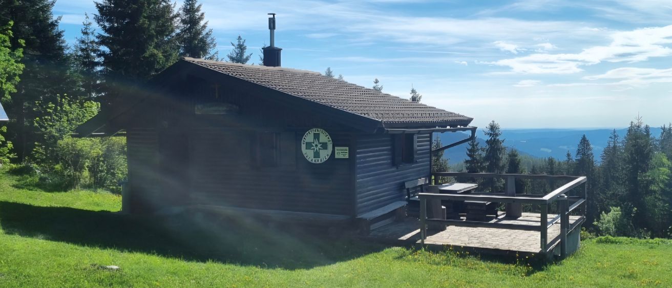 Ein kleines Holzhaus mit Schornstein und grünem Schild steht auf einem grünen Hügel mit Blick auf das Meer.