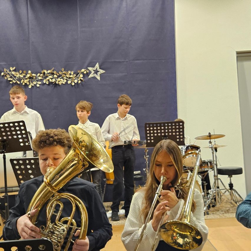 Eine Gruppe junger Musiker in einem Raum spielt Blechblasinstrumente. Ein Junge spielt Tuba, ein Mädchen spielt Posaune. Dahinter spielen andere Flöten. Musikstände und Trommeln sind aufgestellt.