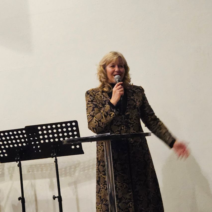 Eine Frau in einem gemusterten Kleid steht an einem Rednerpult, hält ein Mikrofon und lächelt. Hinter ihr befindet sich eine weiße Wand und ein Notenständer mit zwei Musikblättern.