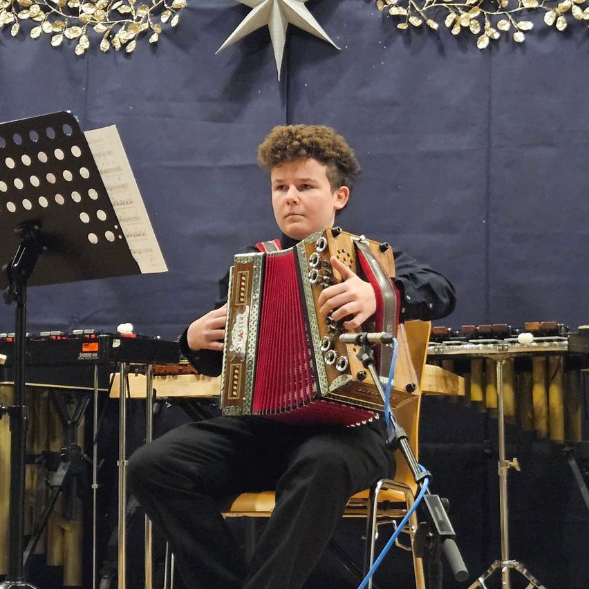 Ein junger Mann spielt auf der Bühne ein Akkordeon, mit einem Notenständer und verschiedenen Instrumenten im Hintergrund.