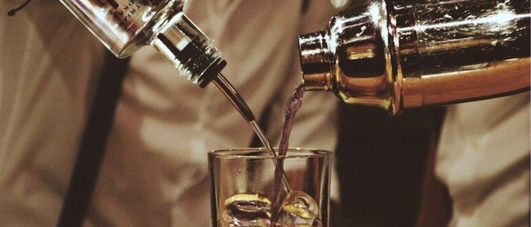 Ein Barkeeper gießt einen Cocktail in ein Glas mit Eiswürfeln auf einem Holztisch.