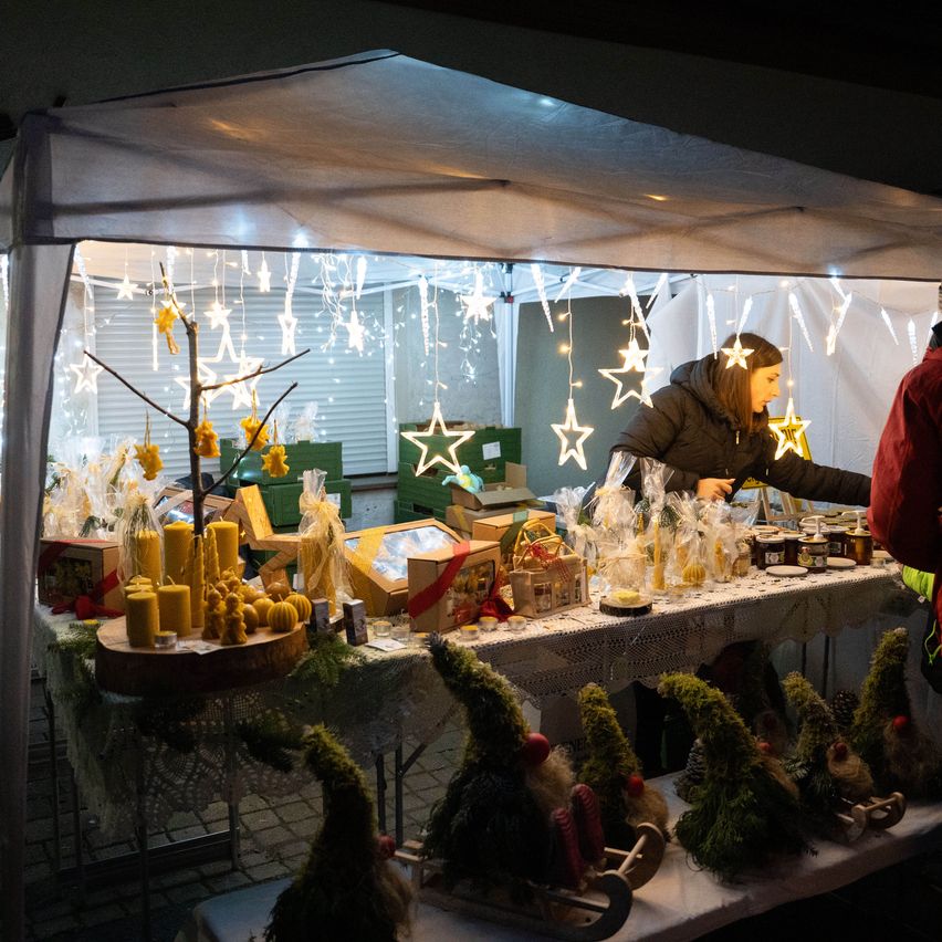 Eine Frau steht in einem Zelt und ordnet Gegenstände auf einem Tisch an, während zwei Personen außerhalb des Zeltes stehen. Der Tisch ist mit verschiedenen Produkten gefüllt, darunter Kerzen, Schachteln und Miniaturfiguren. Das Zelt ist mit Lichterketten und Sternen dekoriert.
