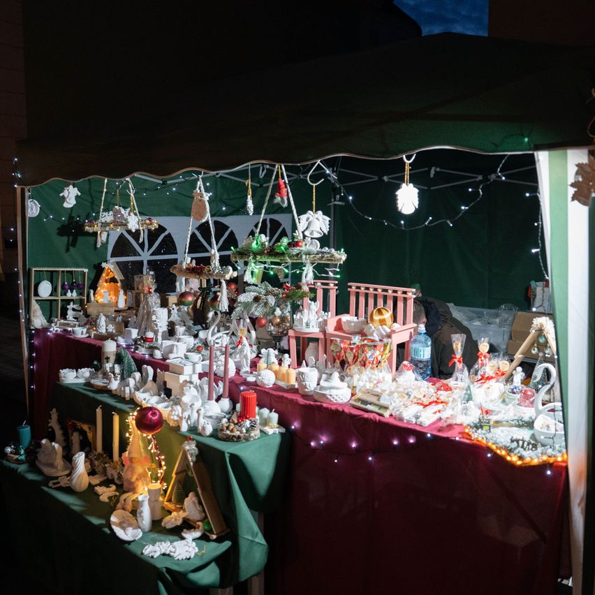 Ein Weihnachtsmarkt-Display unter einem Zelt mit verschiedenen Ornamenten, Figuren und Kerzen auf grünen und roten Tischdecken, beleuchtet von Lichtern.