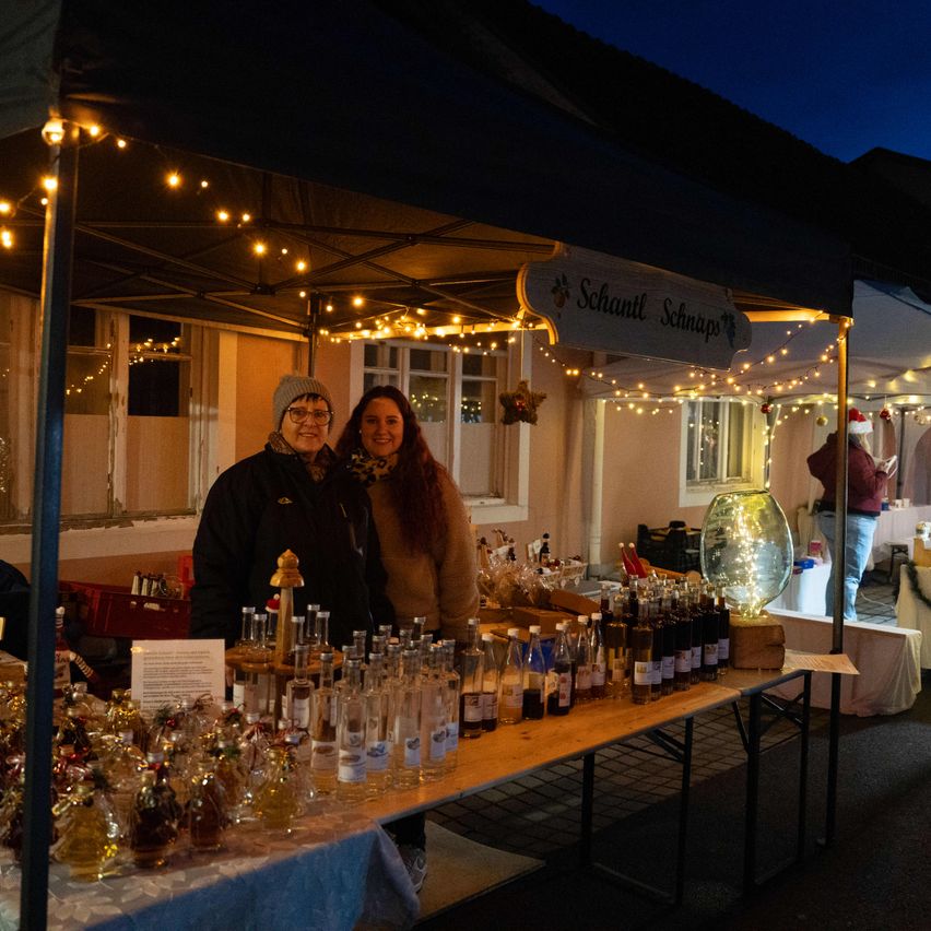 Zwei Frauen stehen unter einem Zelt mit Lichterketten und einem Schild mit der Aufschrift 'Schantl Schnapps'. Flaschen mit Schnapps stehen auf einem Tisch, mit vielen weiteren Flaschen dahinter. Eine weitere Person steht im Hintergrund.