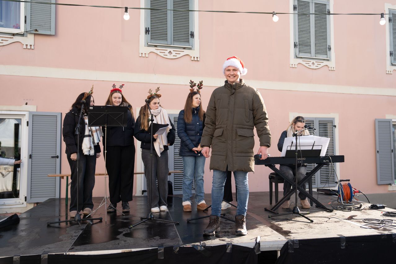 Eine Gruppe von Frauen, gekleidet in Weihnachtsmützen und Schals, steht auf der Bühne. Eine Frau, die lächelt, steht vor einem Mikrofon. Hinter ihr halten andere Frauen Notenständer. Eine weitere Frau spielt Klavier. Die Bühne befindet sich vor einem rosa Gebäude.
