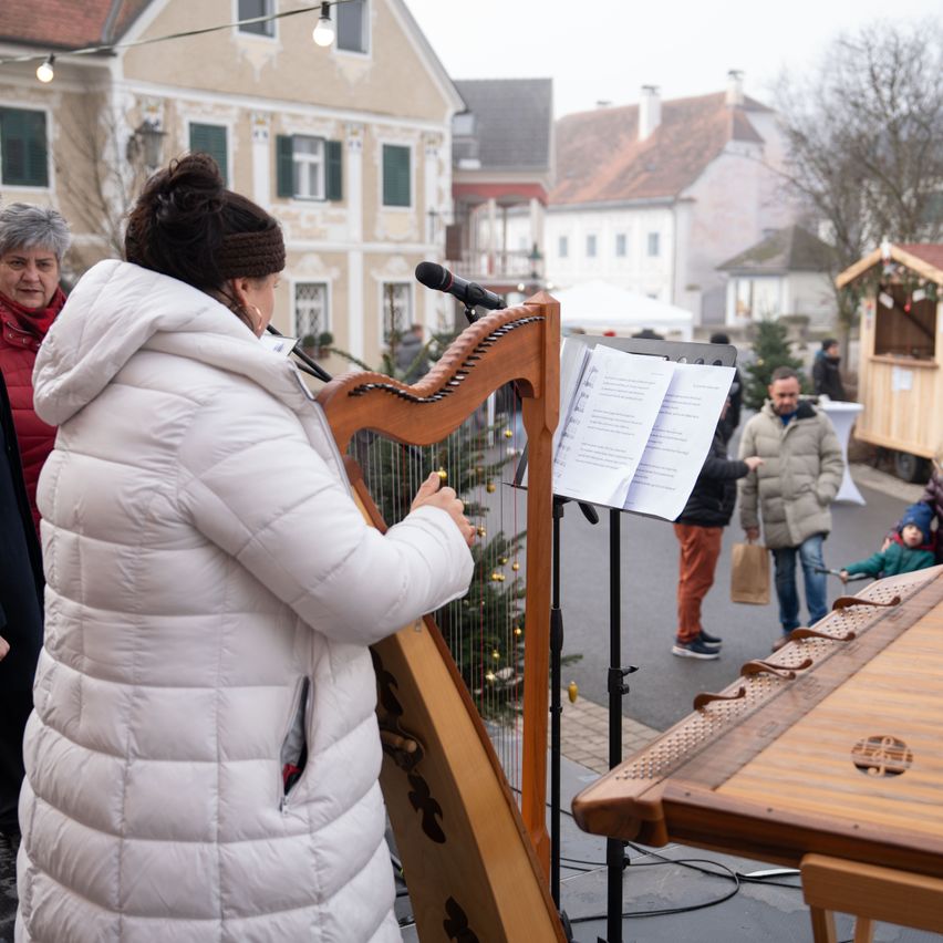Eine Frau in einem weißen, gepolsterten Mantel spielt Harfe vor einem Mikrofon. Hinter ihr steht ein geschmückter Weihnachtsbaum. Andere Leute stehen in der Nähe, einer von ihnen hält ein Kind.