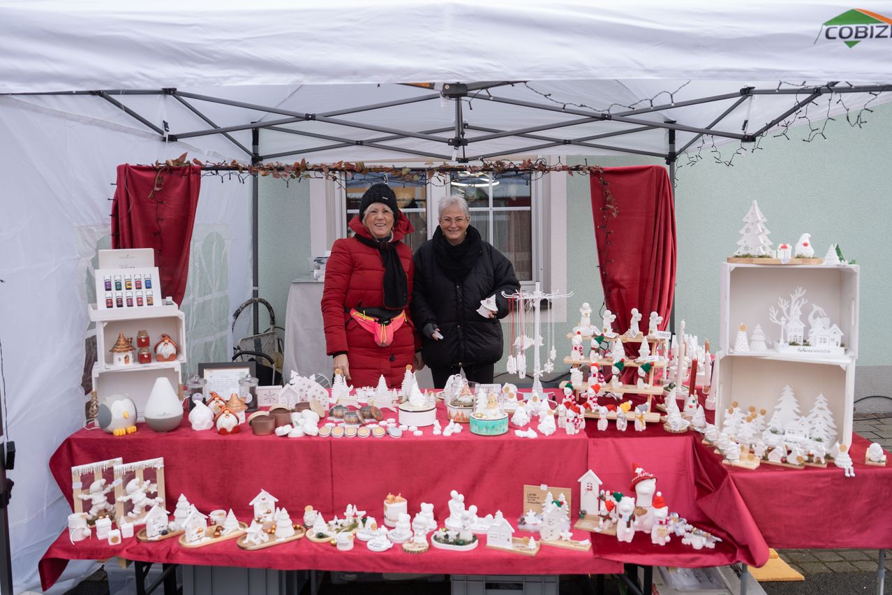 Zwei Frauen stehen hinter einem Tisch mit einem roten Tuch und präsentieren Weihnachtsdekorationen. Der Tisch ist gefüllt mit kleinen Keramikhäusern, Figuren und Ornamenten. Sie lächeln und scheinen ihre Zeit zu genießen.