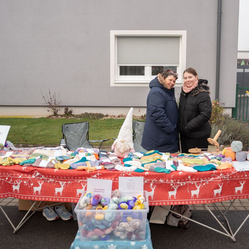 Zwei Frauen stehen hinter einem Tisch mit verschiedenen gestrickten Artikeln und Garn, wahrscheinlich auf einem Handwerksmarkt. Der Tisch ist mit einer roten Tischdecke mit weißen Hirschdesigns bedeckt. In der Nähe befinden sich Stühle, ein Aufbewahrungskasten und eine Spinnwirtel.